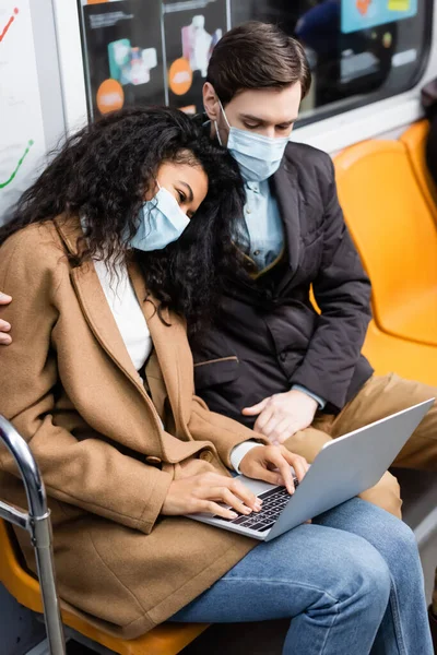Afro-américaine en masque médical à l'aide d'un ordinateur portable près du petit ami dans le métro — Photo de stock