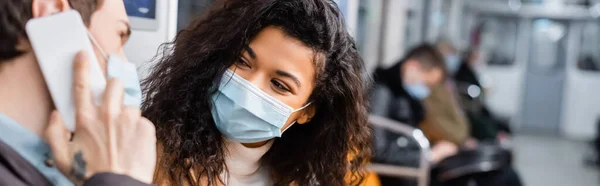 African american woman in medical mask looking at man talking on smartphone on blurred foreground, banner — Stock Photo