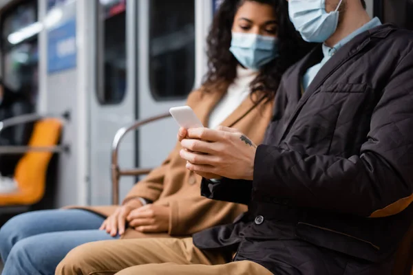 Homem usando smartphone no metrô perto de mulher americana africana em máscara médica — Fotografia de Stock