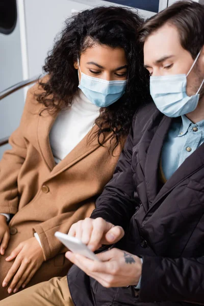 Чоловік у медичній масці за допомогою смартфона біля афроамериканської жінки в метро — стокове фото