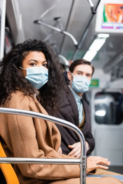 Mulher americana africana em máscara médica olhando para a câmera perto do homem no metrô em fundo borrado — Fotografia de Stock