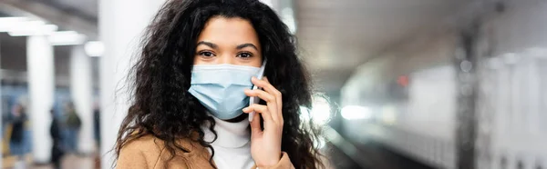 Mujer afroamericana en máscara médica hablando en el teléfono inteligente en metro, pancarta - foto de stock