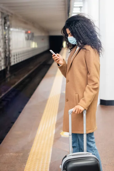 Femme afro-américaine en masque médical debout avec des bagages et en utilisant un smartphone dans le métro — Photo de stock