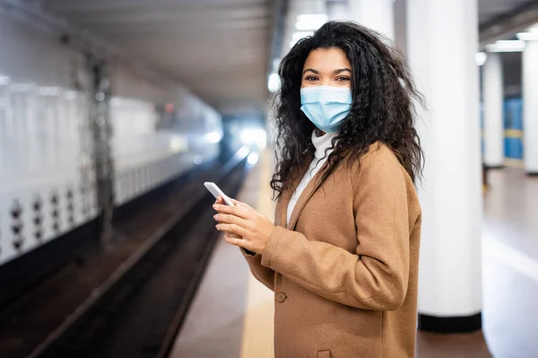 Frisée afro-américaine en masque médical en utilisant un smartphone dans le métro — Photo de stock