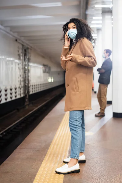 Pleine longueur de jeune femme afro-américaine en masque médical parlant sur smartphone dans le métro — Photo de stock