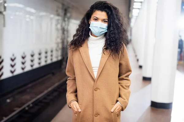 Femme afro-américaine bouclée en masque médical debout avec les mains dans les poches dans le métro — Photo de stock