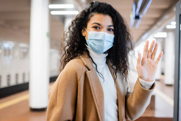 Lockige Afroamerikanerin mit medizinischer Maske winkt in der U-Bahn — Stockfoto