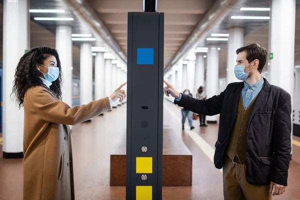 Young african american woman and man in medical masks pointing with fingers at information stand in subway — Stock Photo
