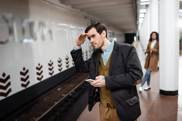 Чоловік тримає смартфон і дивиться далеко від афроамериканської жінки в метро на розмитому фоні — стокове фото