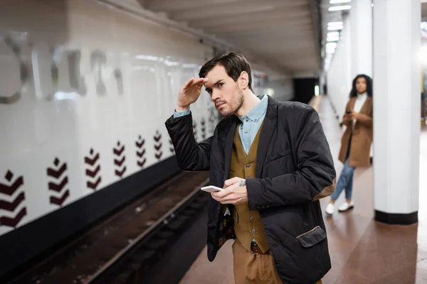 Homem segurando smartphone e olhando para longe perto de mulher americana africana no metrô no fundo borrado — Fotografia de Stock