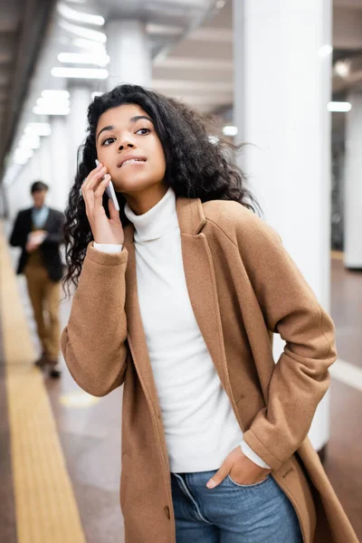 Mulher americana africana mordendo lábios enquanto fala no smartphone e olhando para longe no metrô no fundo borrado — Fotografia de Stock