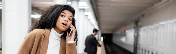 Mujer afroamericana sorprendida hablando en teléfono inteligente y mirando hacia otro lado en el metro sobre fondo borroso, pancarta - foto de stock