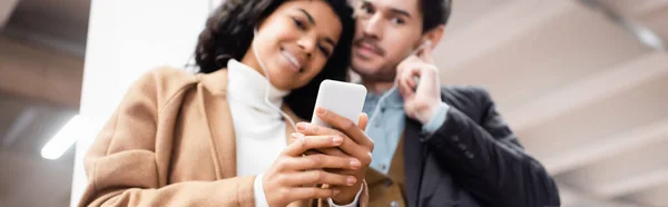 Baixo ângulo de visão do casal multicultural com smartphone ouvindo música em fones de ouvido no metrô em fundo embaçado, banner — Fotografia de Stock