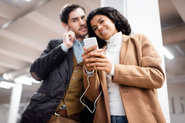 Vista en ángulo bajo de pareja multicultural con smartphone escuchando música en auriculares en metro sobre fondo borroso - foto de stock
