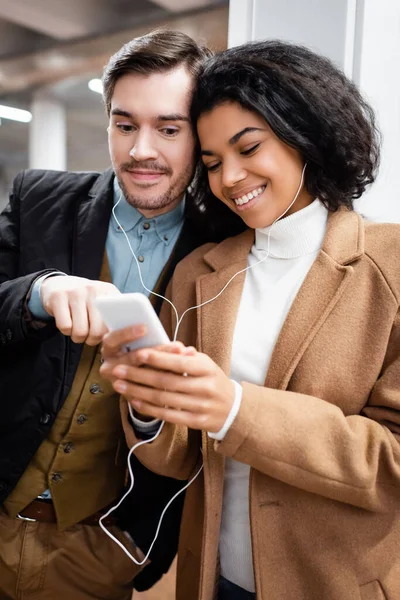 Feliz pareja multicultural mirando el teléfono inteligente y escuchando música en los auriculares en el metro - foto de stock