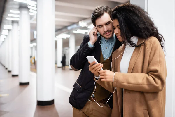 Multikulturelles Paar schaut auf Smartphone, während es in der U-Bahn über Kopfhörer Musik hört — Stockfoto