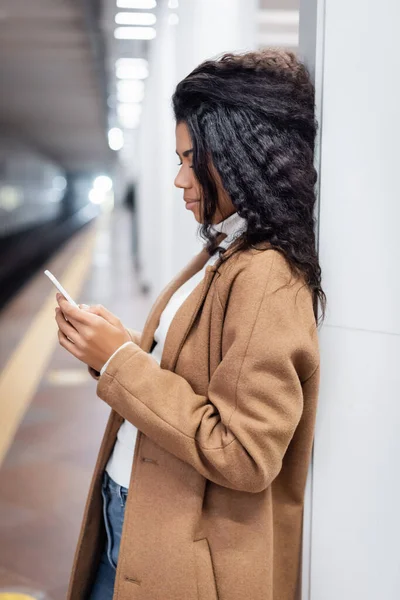 Vista lateral de la mujer afroamericana rizada usando teléfono inteligente en el metro - foto de stock