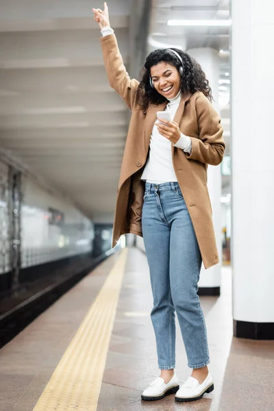 Piena lunghezza della donna afroamericana eccitata in cuffie wireless utilizzando smartphone durante l'ascolto di musica in metropolitana — Foto stock