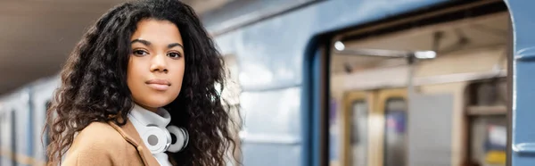 Mujer afroamericana joven en auriculares inalámbricos mirando la cámara en el metro, pancarta - foto de stock