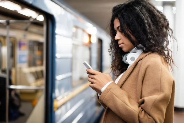 Vista lateral da mulher americana africana em fones de ouvido sem fio usando smartphone no metrô — Fotografia de Stock