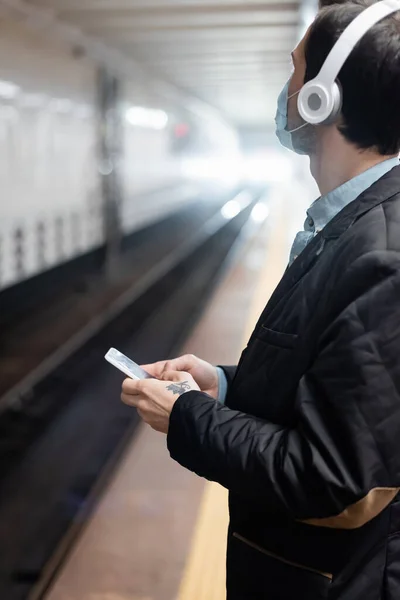 Homem tatuado em máscara médica e fones de ouvido segurando smartphone no metrô — Fotografia de Stock