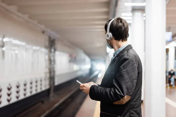 Homem tatuado em máscara médica e fones de ouvido sem fio segurando smartphone no metrô — Fotografia de Stock