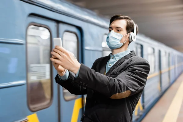 Uomo in maschera medica e cuffie senza fili scattare selfie su smartphone in metropolitana — Foto stock