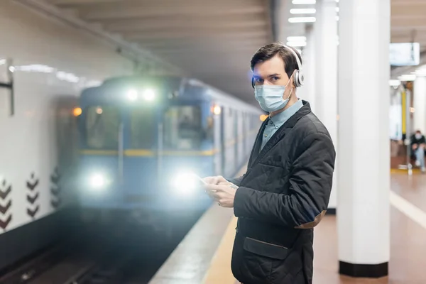 Uomo in maschera medica e cuffie wireless con smartphone in metropolitana — Foto stock