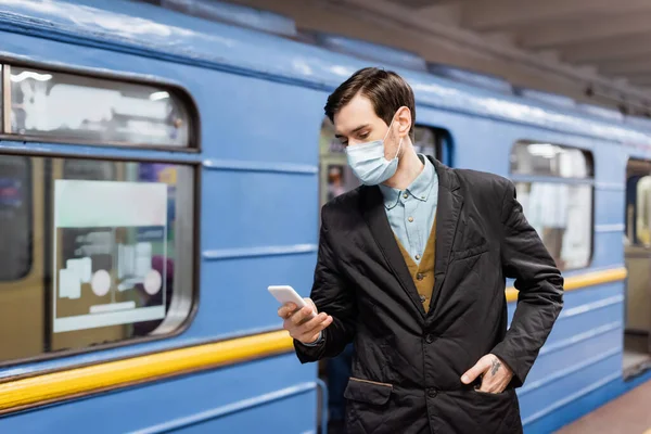 Homem tatuado em máscara médica segurando smartphone e de pé com a mão no bolso perto do vagão do metro — Fotografia de Stock