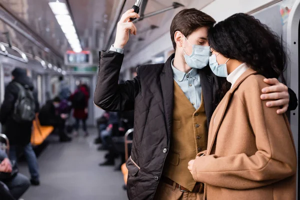 Hombre en máscara médica abrazando afro-americana novia en vagón de metro con la gente sobre fondo borroso - foto de stock