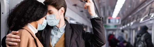 Hombre en máscara médica abrazando a la novia afroamericana en vagón de metro con la gente sobre fondo borroso, pancarta - foto de stock