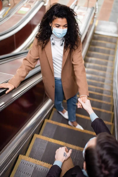 Vue grand angle de la femme afro-américaine dans le masque médical tenant la main avec petit ami sur l'escalator dans le métro — Photo de stock