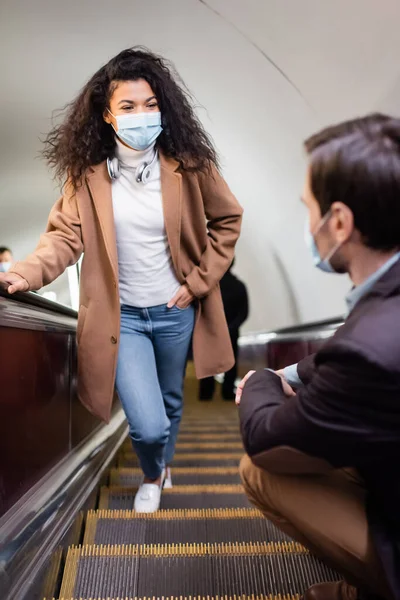 Mujer afroamericana en máscara médica de pie con la mano en el bolsillo en la escalera mecánica cerca del hombre en primer plano borrosa - foto de stock