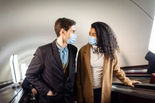 Interracial couple in medical masks looking at each other on escalator in subway — Stock Photo