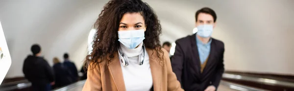 Donna afroamericana in maschera medica vicino all'uomo sulla scala mobile su sfondo sfocato, striscione — Foto stock