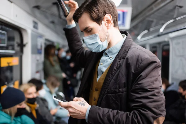 Татуированный человек в медицинской маске с помощью смартфона, стоя в вагоне метро — стоковое фото