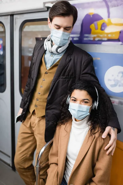 Hombre en auriculares abrazando a mujer afroamericana en máscara médica en metro - foto de stock