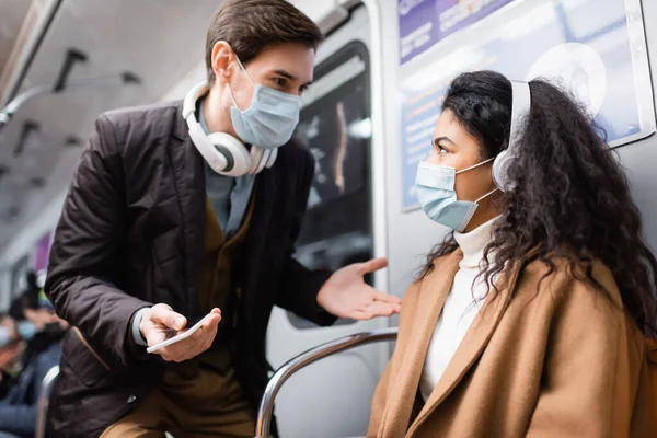 Homem em fones de ouvido segurando smartphone e gestos perto de mulher americana africana em máscara médica no metrô — Fotografia de Stock