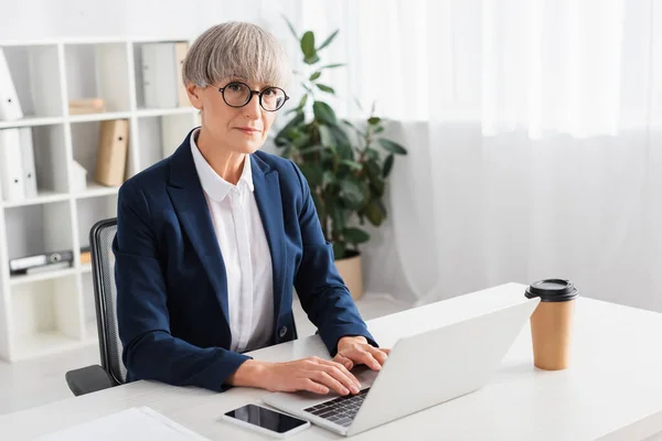 Leader del team di successo digitando sulla tastiera del computer portatile vicino smartphone con schermo bianco e caffè per andare — Foto stock