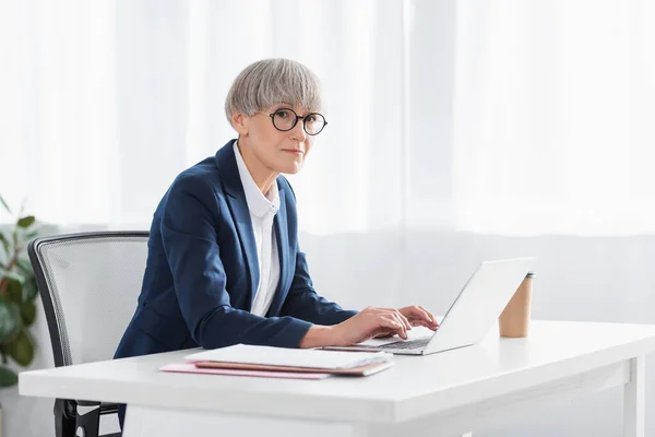 Exitoso líder de equipo escribiendo en el teclado del ordenador portátil cerca de café para ir en el escritorio - foto de stock