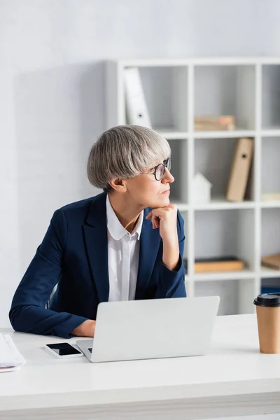 Pensativo líder del equipo en gafas mirando hacia otro lado cerca de gadgets y café para ir en el escritorio - foto de stock