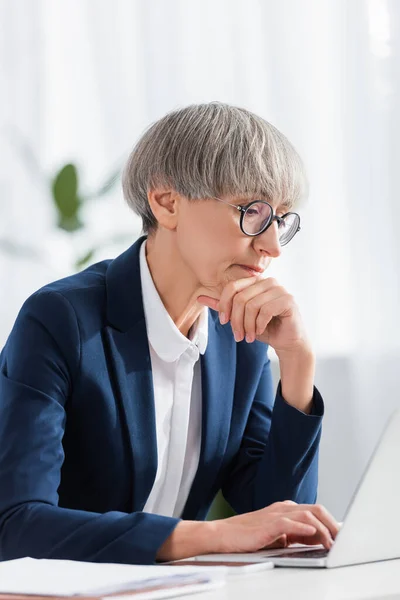 Líder de equipe pensativo em óculos olhando para laptop no escritório — Fotografia de Stock