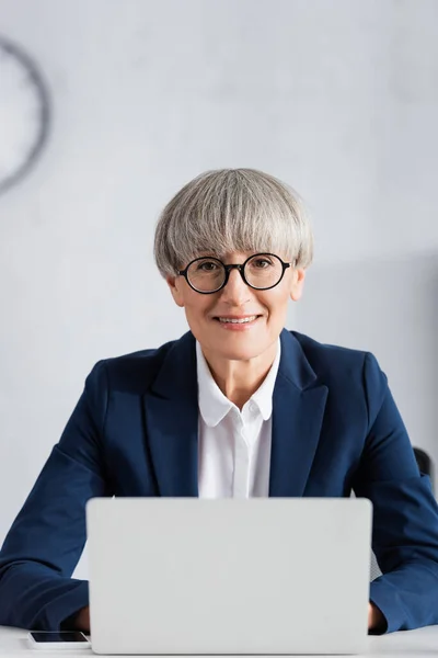 Alegre líder del equipo en gafas sonriendo cerca del ordenador portátil sobre fondo borroso - foto de stock