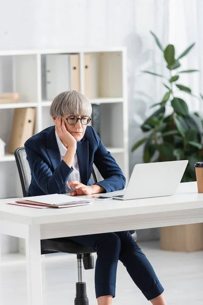 Aburrido líder del equipo en gafas mirando portátil en el escritorio en la oficina moderna - foto de stock