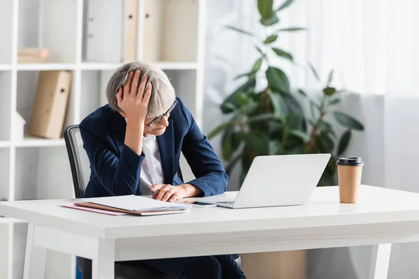 Líder de equipe cansado em óculos olhando para laptop na mesa no escritório moderno — Fotografia de Stock