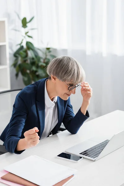 Middle aged businesswoman in glasses with clenched fist suffering from headache in office — Stock Photo