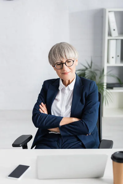 Mature team leader with crossed arms looking at laptop on desk — Stock Photo