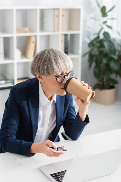 Leader del team di mezza età in bicchieri di caffè per andare mentre si utilizza smartphone vicino al computer portatile sulla scrivania — Foto stock