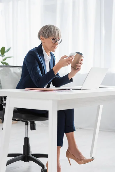 Middle aged team leader in glasses holding coffee to go while using smartphone near laptop on desk — Stock Photo