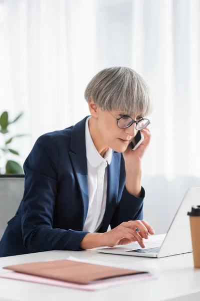 Geschäftsfrau mittleren Alters mit Brille spricht auf Smartphone, während sie Laptop benutzt — Stockfoto